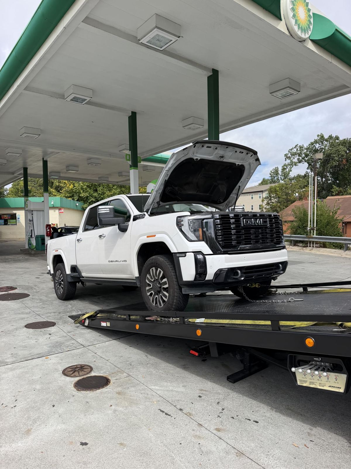 Flatbed tow truck loading a white GMC Sierra HD with its hood up at a gas station